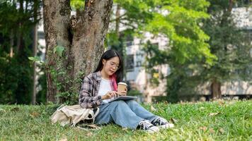 uma jovem ásia mulher senta debaixo uma árvore dentro uma campus parque, segurando uma café copo e uma tábua. foto