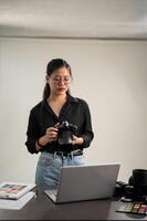 a elegante ásia fêmea fotógrafo dentro uma Preto camisa com dela dslr Câmera é em pé dentro dela estúdio foto