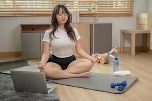 uma calma, relaxado jovem ásia mulher dentro roupa de esporte é meditando em uma ioga esteira dentro a vivo sala. foto