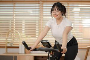 uma Forte ásia mulher dentro roupa de esporte é exercício em uma ginástica bicicleta, fazendo interior cardio às lar. foto
