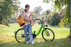 uma lindo ásia mulher é usando dela Smartphone enquanto caminhando com dela bicicleta dentro uma ensolarado verde parque. foto