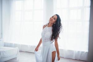 satisfeito e livre. Mulher bonita em um vestido branco fica em um quarto branco com a luz do dia entrando pelas janelas foto