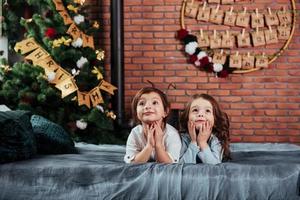 em antecipação a presentes. o que elas duas garotas alegres deitadas na cama com decorações de ano novo e árvore de Natal foto