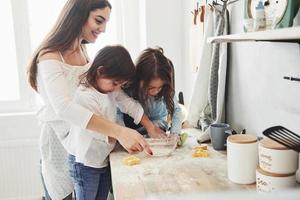vista lateral. mãe e duas meninas na cozinha estão aprendendo a cozinhar uma boa comida com farinha foto