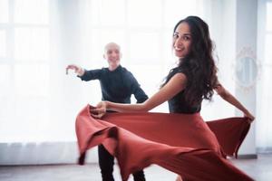 gostando de fazer a performance. Mulher jovem e bonita em roupas vermelhas e pretas dançando com um cara careca na sala branca foto