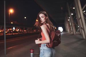 uma linda jovem com uma mochila atrás dos ombros está na rua perto de um aeroporto ou estação ferroviária em uma noite quente de verão. ela acabou de chegar e espera por um táxi ou seus amigos foto