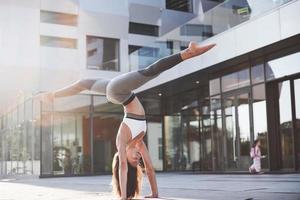 manhã ensolarada de verão. jovem atlética fazendo parada de mãos na rua do parque da cidade, entre modernos edifícios urbanos. exercício ao ar livre estilo de vida saudável foto