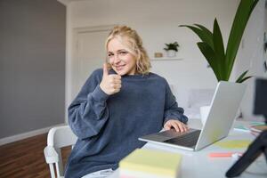 retrato do sorridente jovem loiro mulher, menina aluna estudando a partir de lar, usando conectados Academia, fazendo curso dentro Internet, usando computador portátil, mostrando polegares acima. conceito do distância Aprendendo foto