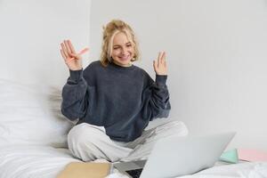 retrato do jovem feliz mulher, conecta para bater papo, usando computador portátil, acenando mão às Câmera, dizendo Olá para alguém on-line, sentado em cama, estudo, e-learning ou trabalhando a partir de casa foto