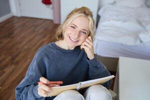 retrato do sorridente, lindo jovem sincero mulher, sentado dentro quarto com caneta e caderno, escrevendo dentro Diário, fazer notas, olhando feliz às Câmera foto