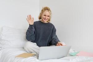 retrato do jovem feliz mulher, conecta para bater papo, usando computador portátil, acenando mão às Câmera, dizendo Olá para alguém on-line, sentado em cama, estudo, e-learning ou trabalhando a partir de casa foto