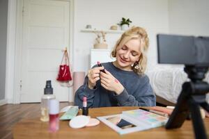 retrato do lindo jovem mulher, conteúdo O Criador para social meios de comunicação, sentado dentro frente do digital Câmera, gravação sobre inventar, mostrando batom amostras em dela pele foto