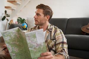 retrato do jovem homem, viajante, sentado embalagem mala, olhando às viagem mapa, escolhendo feriado destino, estudando a rota em período de férias foto