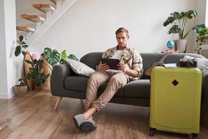 imagem do jovem homem com tábua, sentado dentro vivo quarto com mala, leitura, estudando a rota, preparando para uma período de férias foto
