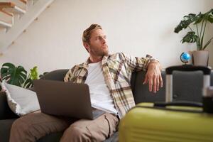 feliz homem reserva feriado conectados em computador portátil, pagando para período de férias em viagem agência local na rede Internet, comprando voar bilhetes dentro Internet, sentado dentro vivo quarto com mala de viagem foto