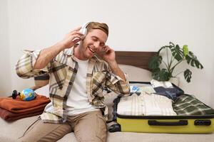 bonito loiro homem dentro fones de ouvido, senta em cama com mala de viagem e roupas, escuta para música dentro sem fio fones de ouvido enquanto embalagem para uma feriado, indo em viagem foto