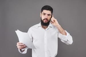 homem de negócios com barba na camisa branca segurando documentos e o telefone em fundo cinza