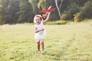 menina feliz correndo no campo com o avião de brinquedo vermelho nas mãos. árvores no fundo foto