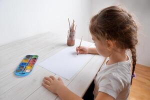 pequeno menina tintas com água cores em branco papel. conceito do criatividade, arte, infância, imaginação, e artístico expressão foto