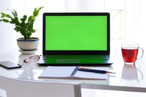 zombar acima, computador portátil com verde tela, conceito casa trabalhar, distância Educação. caderno, caneta, Smartphone, copos, flor, copo do chá em a mesa dentro frente do janela. foto