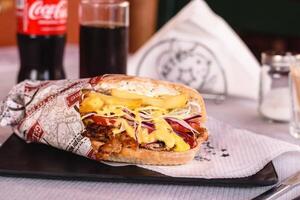 giros carne de porco com queijo, tomate, molho, fritas às grego feito à mão pastelaria pitsopsomo com sésamo servido em a Preto prato em branco fundo dentro grego taberna. Coca Cola em fundo. seletivo foco foto