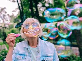 mulher elegante sênior com cabelos grisalhos e óculos azuis e jaqueta jeans, soprando bolhas ao ar livre. feriados, festa, anti-idade, conceito divertido foto