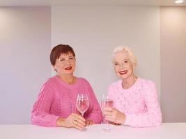 duas elegantes mulheres sêniors em suéteres cor de rosa bebendo vinho rosé na cozinha moderna fofocando. amizade, conversa, fofoca, evento, relacionamentos, notícias, conceito de família foto