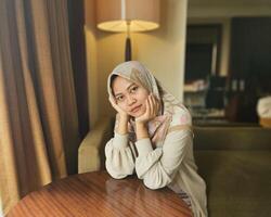 ásia muçulmano mulher vestindo hijab sentado pensativamente enquanto apoiando queixo e cabeça, olhando às Câmera com borrado fundo foto