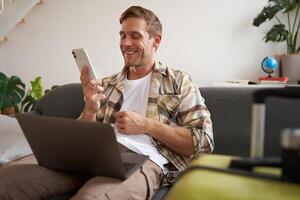 sorridente bonito homem indo em feriado, fazer uma telefone chamar, segurando computador portátil, preparado mala de viagem para uma viagem no exterior foto