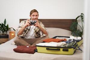 alegre jovem homem leva uma cenário em digital Câmera, senta em cama, vai em feriado, pacotes roupas para férias, turista animado antes indo em viagem, viajando foto