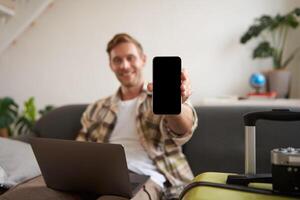 retrato do bonito jovem homem, mostrando Smartphone tela, recomendando Móvel aplicativo para viajando ou reserva on-line, sentado com computador portátil e mala de viagem foto