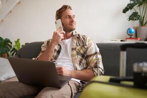 bonito sorridente homem com computador portátil, fazer uma telefone chamar, olhando a parte, de lado enquanto esperando em a Telefone, confirma reserva conectados foto