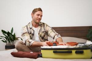 retrato do sorridente bonito homem, senta em cama, coloca roupas dentro mala, vai em férias, sonhos sobre feriado, livros uma viagem com viagem agência e pacotes coisas foto