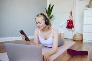 retrato do feliz, sorridente desportivo mulher, fazendo exercite-se às lar, usando computador portátil para Assistir exercite-se s dentro fones de ouvido, verificação Esportes aplicativo em Móvel telefone foto