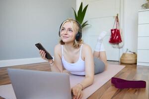 retrato do feliz, sorridente desportivo mulher, fazendo exercite-se às lar, usando computador portátil para Assistir exercite-se s dentro fones de ouvido, verificação Esportes aplicativo em Móvel telefone foto