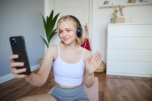 retrato do jovem mulher, ginástica instrutor fazendo conectados exercite-se sessão em Smartphone, acenando mão às Câmera e dizendo olá, sentado em borracha esteira dentro uma quarto foto