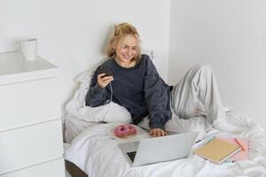 estilo de vida e pessoas conceito. jovem lindo mulher, ficando às lar, deitado dentro cama com computador portátil e Smartphone, comendo rosquinha, desfrutando livre tempo, gastos final de semana às lar, assistindo filme conectados foto