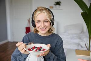 retrato do sorridente fofa mulher dentro fones de ouvido, comendo dela café da manhã e assistindo em computador portátil, olhando às tela com feliz face foto