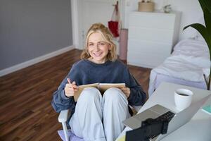 retrato do jovem mulher lendo dela notas em Diário, sentado às casa em cadeira, sorridente e olhando feliz, fazendo dever de casa foto