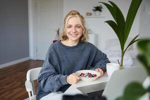 retrato do jovem mulher comendo saudável refeição dentro uma sala, assistindo s em computador portátil, tendo almoço dentro frente do computador, sorridente e olhando feliz às Câmera foto