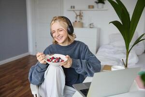 imagem do feliz mulher sentado dentro uma sala, assistindo interessante televisão mostrar ou filme em computador portátil, usando gritando serviço, vestindo fones de ouvido, comendo sobremesa e bebendo chá foto