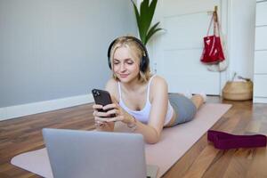 retrato do jovem mulher dar certo, assistindo exercício s em computador portátil dentro fones de ouvido, deitado em borracha esteira com Móvel telefone e sorridente foto