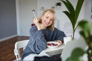 retrato do sorridente loiro jovem mulher, comendo dentro frente do computador portátil, assistindo s conectados enquanto tendo café da manhã, desfrutando sobremesa, sentado dentro quarto foto