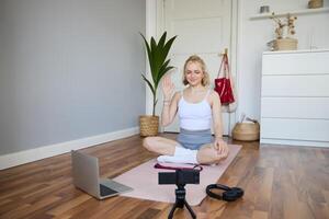 retrato do jovem fêmea atleta, ginástica treinador gravação vlog, Treinamento sessão em digital Câmera, sentado dentro uma quarto em borracha ioga esteira, mostrando exercícios foto