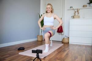 retrato do jovem Atlético mulher, vlogger fazer uma sobre casa dar certo, ginástica instrutor tiroteio quão para Faz perna exercícios, usando elástico resistência banda, usando ioga esteira foto