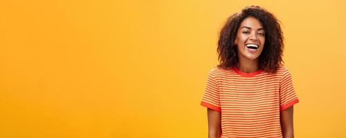 cintura para cima tiro do extrovertido feliz encantador pele escura fêmea com encaracolado Penteado rindo alegremente posando dentro listrado na moda camiseta sobre laranja fundo desfrutando agradável casual conversação foto