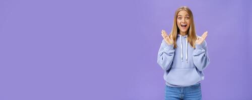 retrato do divertido animado e entretido feliz encantador mulher com justo cabelo dentro na moda moletom com capuz aperto mãos a partir de deleite e surpresa sorridente amplamente alegremente posando contra roxa fundo foto