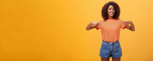 Ei escolher mim Eu seu menina olhando para. retrato do encantador de aparência amigável ambicioso pele escura fêmea com afro Penteado apontando às peito orgulhosamente e alegre posando contra laranja fundo foto