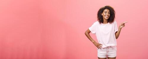 Ei olhar. retrato do fofa calafrio e amigáveis à moda africano americano mulher com afro Penteado segurando mão em cintura apontando certo e sorridente alegremente sobre Rosa fundo foto