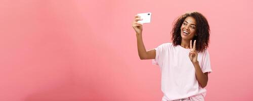 à moda sociável boa aparência pele escura fêmea aluna com encaracolado Penteado puxar mão com Smartphone perto face levando selfie mostrando Paz placa para dispositivo tela enquanto sorridente despreocupado foto